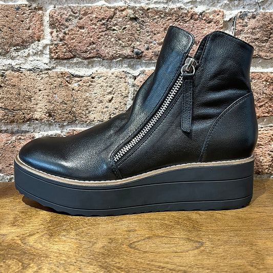 A black leather ankle boot with dual zipper details and a platform sole, displayed against a brick wall backdrop.
