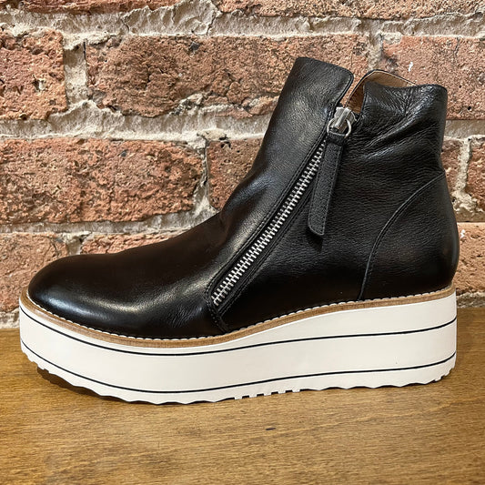 A black leather ankle boot with dual zipper details and a white platform sole, placed on a wooden surface against a brick wall backdrop.