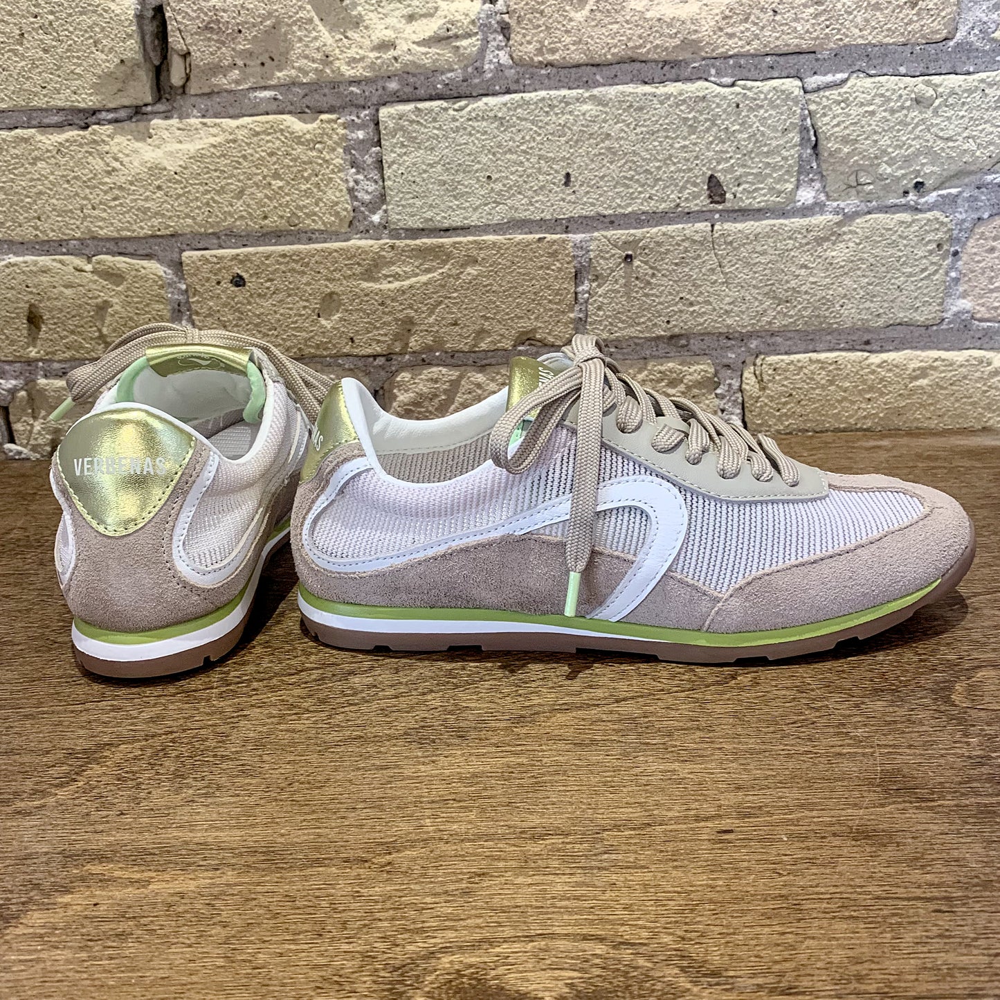 Pair of light gray and white sneakers with green accents on a wooden surface with a brick wall background.