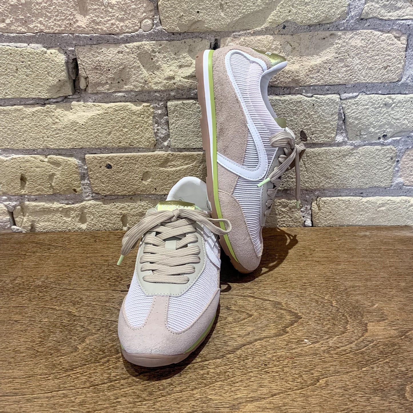 Pair of beige sneakers with white soles on a wooden surface against a brick wall background